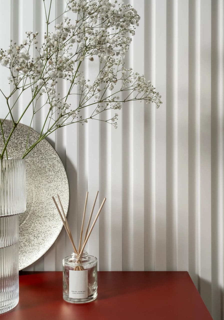 Moldura decorativa blanca en pared con jarrón, flores secas y difusor sobre mesa roja