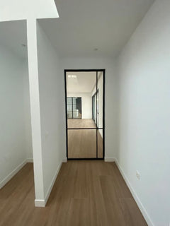 Pasillo moderno con suelo de madera, paredes blancas y puerta de cristal con marco negro.
