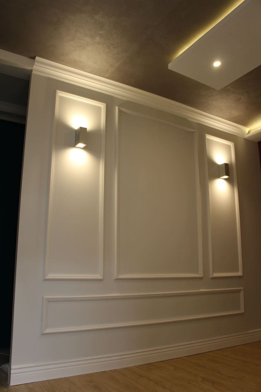 Pared decorada con molduras blancas clásicas y apliques de luz, suelo de madera clara.