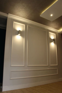 Pared decorada con molduras blancas clásicas y apliques de luz, suelo de madera clara.