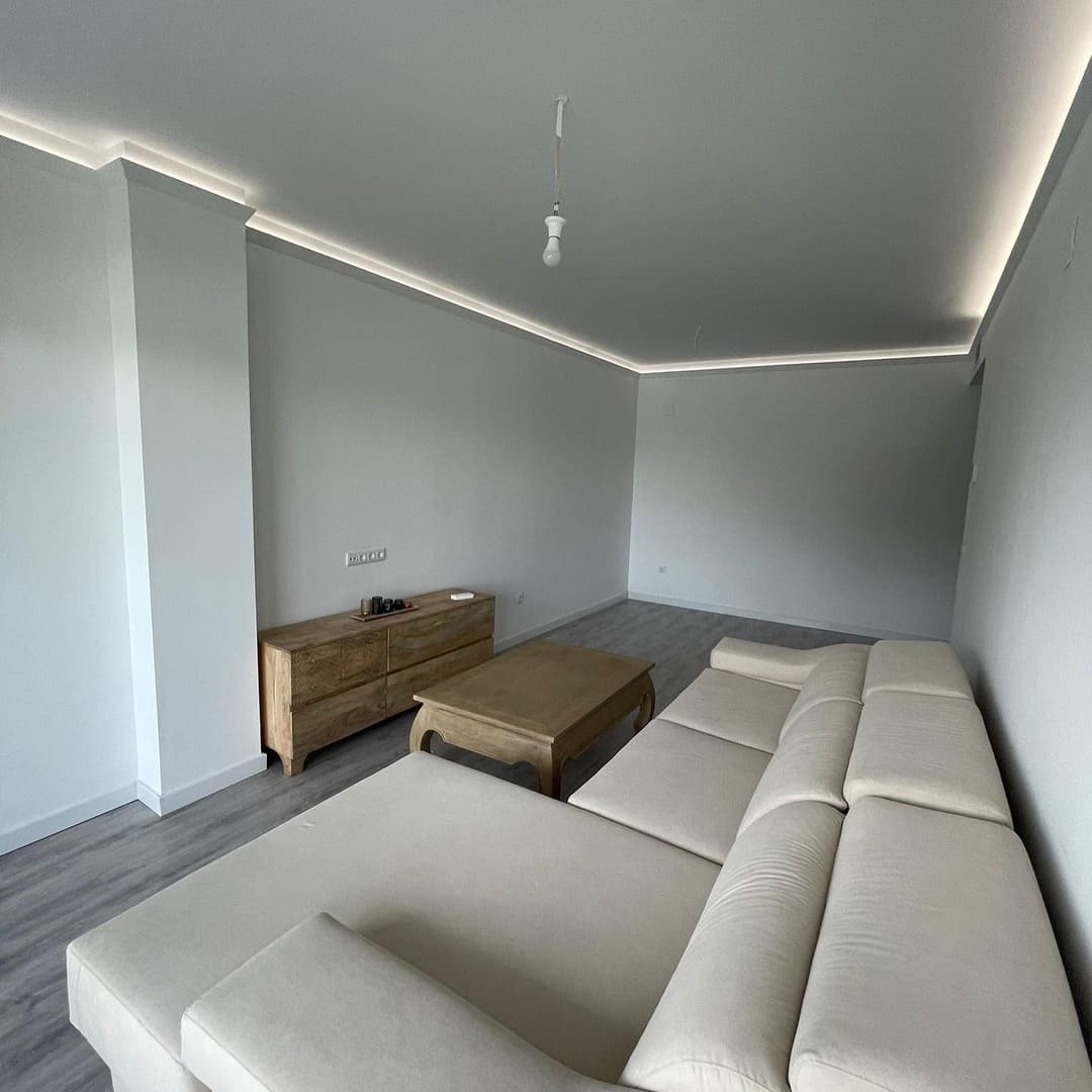 Salón moderno con molduras decorativas, sofá beige y muebles de madera, suelo gris.