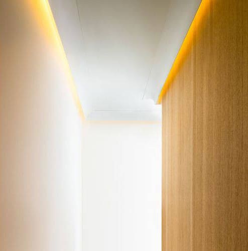 Moldura decorativa de techo con iluminación LED indirecta en pasillo moderno de pared blanca y panel de madera.